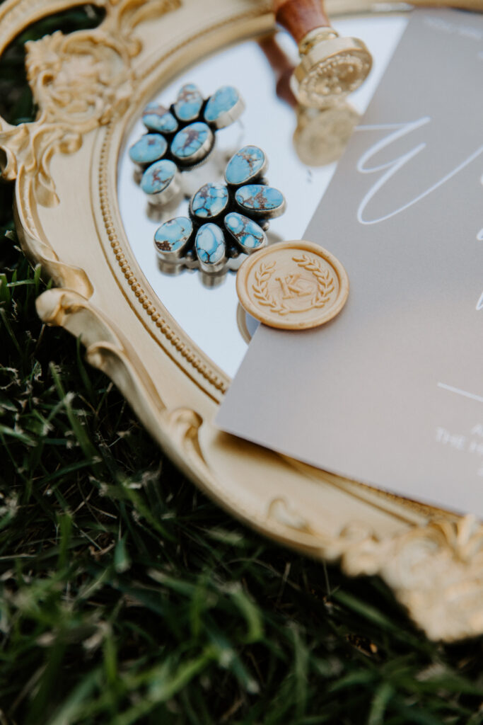 
Elegant wedding detail shot featuring turquoise floral earrings on a vintage gold ornate mirror tray, alongside a wax seal stamp and a wedding invitation, set on lush green grass.