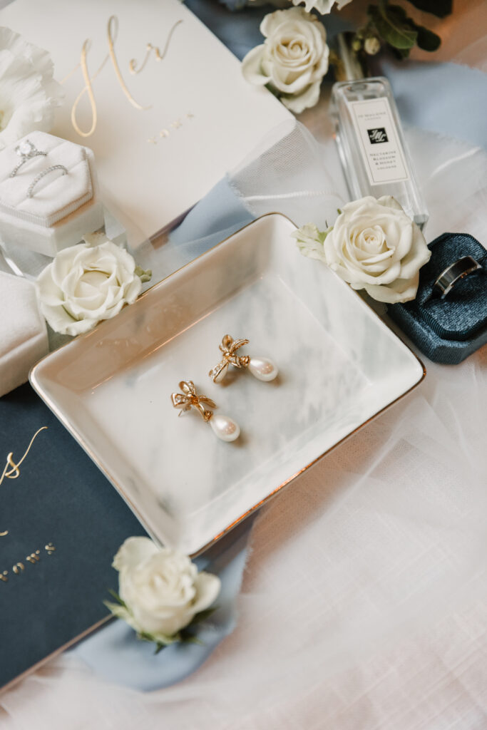 Elegant flat lay wedding preparation scene featuring a pair of pearl earrings with gold bows in a marble dish, white rose accents, Jo Malone perfume, a "Her Vows" book, and white jewelry boxes with rings, all arranged on soft blue and white fabric.