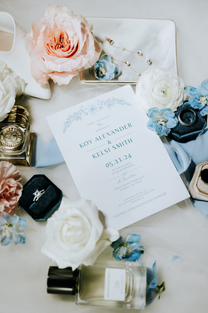 Flat lay wedding detail shot featuring an invitation for Koy Alexander and Kelsi Smith's wedding on May 11, 2024, surrounded by elegant items: a diamond engagement ring in a blue velvet box, a bottle of perfume, pearl jewelry, white and pink roses, blue flowers, and a pair of white bridal shoes, all arranged on a soft, light-colored fabric background.