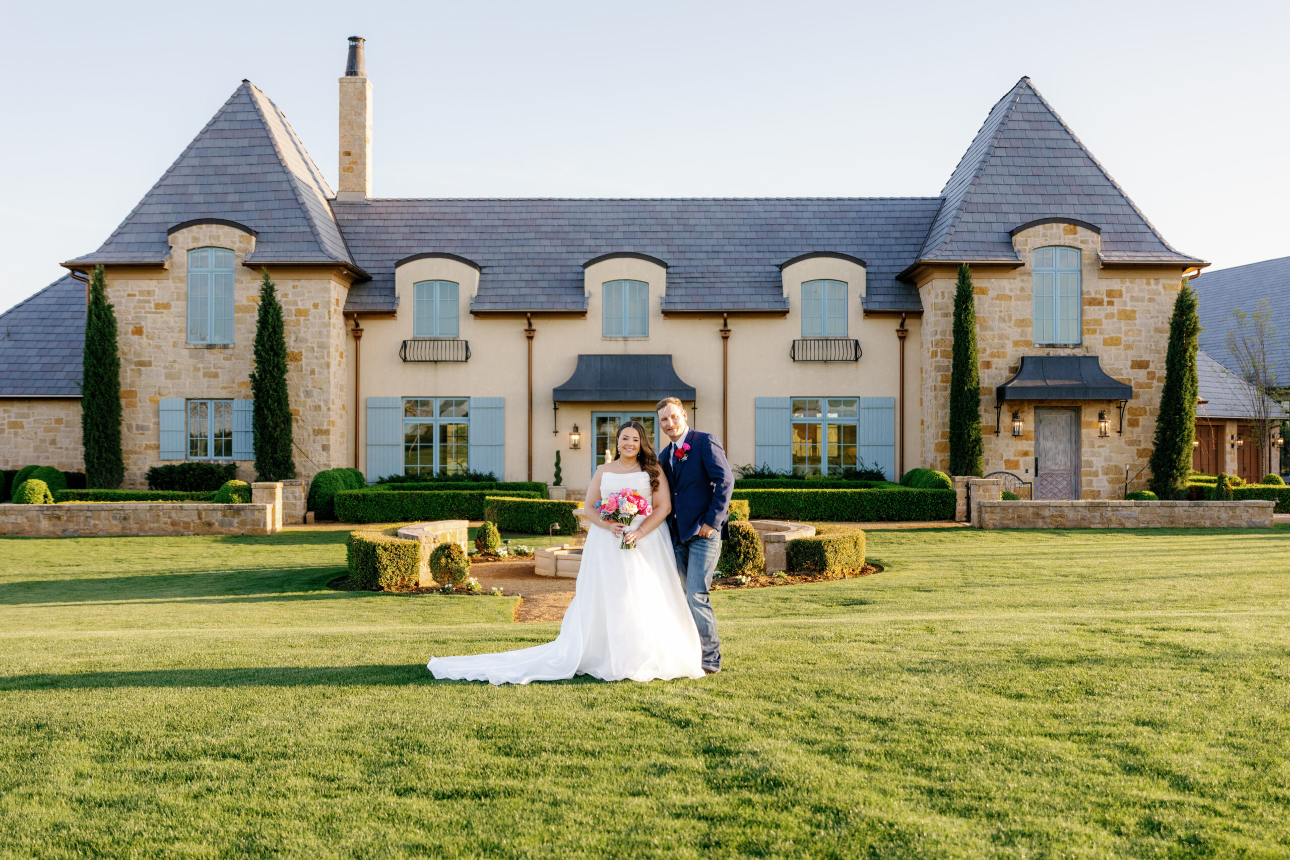 Bride and groom portraits at The Bower standing on a manicured lawn in front of a grand stone estate with symmetrical architecture, soft blue shutters, and warm golden hour light, with the bride holding a colorful bouquet.